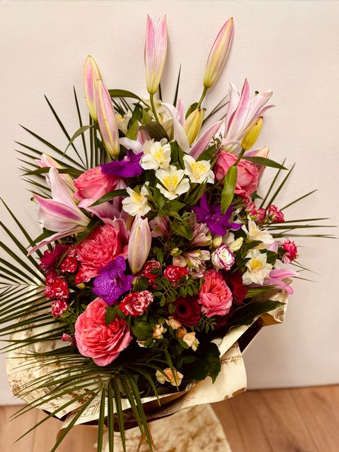 A vibrant floral arrangement featuring pink lilies with tall, closed buds and fully bloomed petals, complemented by large, ruffled pink roses, purple orchids with delicate petals, and smaller white an