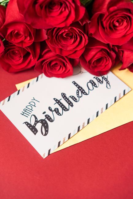 A bouquet of fresh red roses with slightly opened petals and green stems, arranged in a cluster on top of a white and red striped envelope. The arrangement is placed on a red surface, with a 'Happy Bi
