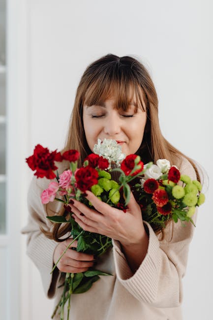 A woman with light brown hair and bangs, dressed in a beige sweater, holds a bouquet of fresh flowers close to her face with her eyes closed and a gentle smile. The bouquet features vibrant red carnat