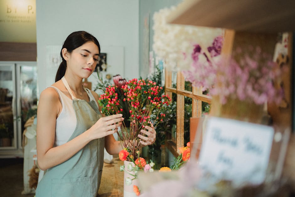 A young woman with dark hair, wearing a light-colored apron over a sleeveless top, is carefully arranging a bouquet of small, vibrant pink and red waxflower blossoms with slender green stems. She stan