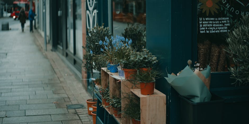 A display of potted plants and flowers arranged on a wooden shelf outside a flower shop, with various greenery and blooming plants in terracotta and ceramic pots, some wrapped in paper. The arrangemen