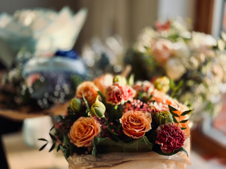 A floral arrangement featuring peach and deep pink roses, complemented by small white filler flowers and green foliage, arranged in a bouquet wrapped in brown kraft paper. The bouquet is placed on a w