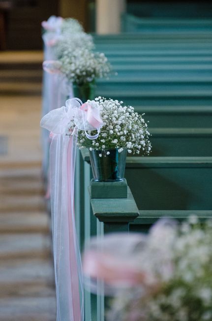 A row of small potted arrangements featuring delicate white baby's breath flowers, placed along a wooden church pew. Each pot is decorated with soft pink and white ribbon bows, adding a gentle touch o