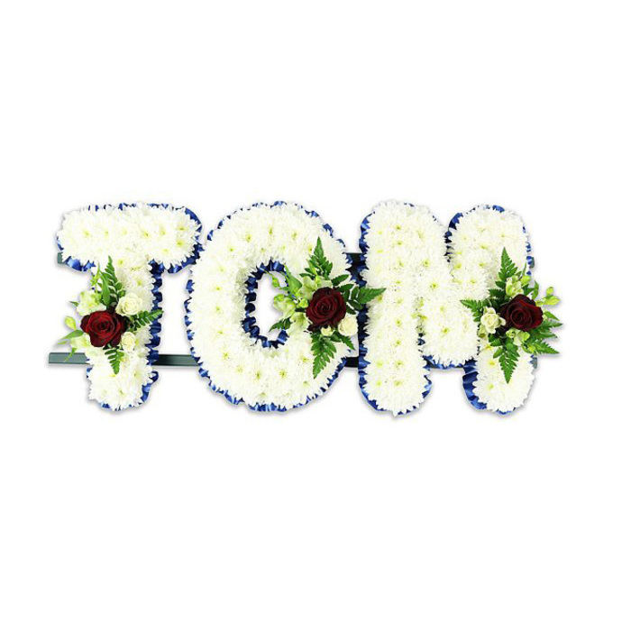 A formal and heartfelt floral tribute that spells the name 'TOM' in bold, three-dimensional letters, each letter carefully formed from a dense, cushion-like mass of small, immaculate white chrysanthemums. The surface of the letters reads soft and textured, a tactile field of petals that absorbs and softens light. Each letter is framed with a vibrant royal blue ribbon along its perimeter, the rich blue acting as a sculptural outline that sharpens the silhouette and adds a dignified accent to the composition. Within the interior of each letter a composed spray is placed: a single dark red rose commands attention at the focal point, its velvety depth evoking love and remembrance, while clusters of creamy white spray roses and slim fern fronds provide gentle contrast and botanical movement. A faint, respectful aroma - faint rose perfume and the vegetal note of fresh greenery - suggests the work of an experienced hand. The letters are mounted on a thin, dark metal support visible at the base, a practical element that does not detract from the solemn beauty above. Set against a plain white background, this arrangement reads cleanly and solemnly, its classic palette of white, royal blue and burgundy lending formality appropriate for chapel display or a civic memorial service. This type of bespoke mourning tribute is the sort of piece local families in Amersham might choose when seeking a personalised expression of sympathy - an arrangement that is both restrained and deeply expressive in its symbolism.