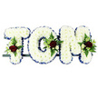 A personalised funeral flower tribute in the image spells the name TOM with grace and clarity, reflecting the kind of thoughtful memorial design often chosen by families in Amersham. The three capital letters are built entirely from crisp white chrysanthemums, tightly packed to form a thick, pillow-like surface with a soft, almost velvety look. Each letter is framed by a royal blue satin ribbon, folded into gentle pleats around the edges so that the blue border neatly defines the outline and brings a traditional, formal character to the design. Against the simple white background, this contrast of pure white petals and strong blue edging stands out beautifully. At three carefully selected points-the upper left of the 'T', the curve of the 'O', and the right-hand side of the 'M'-small, decorative flower sprays introduce richer tones. These clusters feature deep red roses, their velvety petals fully opened, nestled among tiny white or cream spray roses and narrow, trumpet-shaped white flowers reminiscent of freesia. Sprigs of fresh green foliage and delicate fern fronds emerge from behind the blossoms, adding softness, texture, and a sense of natural movement to the structured lettering. The letters are mounted together on a slim, dark bar, arranged in a tidy horizontal row and ready to be placed at the front of a chapel, crematorium, or graveside. The arrangement transforms Tom's name into a floral expression of love, sorrow, and remembrance, surrounding it with gentle colour and the quiet beauty of expert floristry.